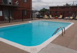 apartment building swimming pool it is filled with clear blue water and surround by a black fence there are empty tan deck chairs along one side