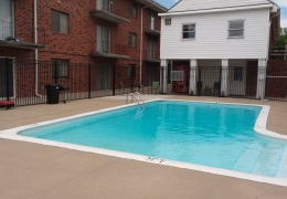 swimming pool filled with clear blue water and surround by a black fence on side of the pool sits a brick apartment building on the other sits a white club house