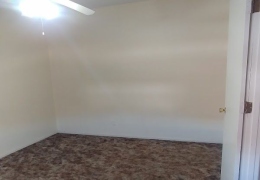 apartment bedroom with dark carpet and cream walls, a ceiling fan and white door. it is empty of furniture and ready for rent