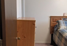 apartment bedroom with light brown carpet and cream walls. there is a brown wood dresser along one wall and a bed with a colored bedspread along the other