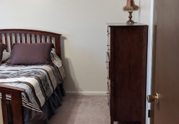 apartment bedroom with light brown carpet and cream walls. there is a dark brown wood dresser along one wall and a bed with a colored bedspread along the other