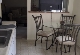 apartment kitchen and dining area the kitchen has white cupboards along one wall and the white linoleum flooring the dining area has a small glass table with 3 black iron chairs around it