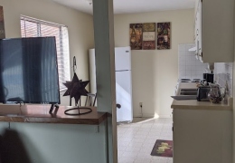 apartment kitchen and dining area. there is a white refrigerator along one wall and cabinets with a sink and stove along the other it has the white linoleum flooring with a dark mat in front of the sink
