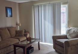 apartment living room with sliding glass door along one wall there is a tan couch and loveseat and coffee table the walls and carpet are cream and there is light streaming in from the door