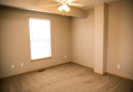 apartment bedroom with light grey carpet and walls, a ceiling fan and light brown closet doors there is light flowing in from the windows and it is empty of furniture and ready for rent