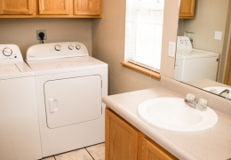 apartment bathroom and laundry room with grey walls, white appliances, light brown cupboards, and white tile floor there is light flowing in from the window in the room