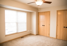 apartment bedroom with light grey carpet and walls, a ceiling fan and light brown closet doors there is light flowing in from the windows and it is empty of furniture and ready for rent