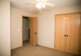 apartment bedroom with light grey carpet and walls, a ceiling fan and light brown closet doors it is empty of furniture and ready to rent