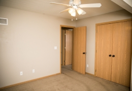 apartment bedroom with light grey carpet and walls, a ceiling fan and light brown closet doors it is empty of furniture and ready to rent