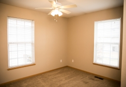 apartment bedroom with light grey carpet and walls, a ceiling fan and light brown closet doors there is light flowing in from the windows and it is empty of furniture and ready for rent
