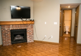 living room of an apartment with wood floors grey walls and fireplace with brick trim there is a side door open to the bathroom with a toilet showing