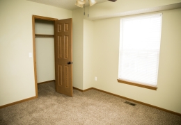 apartment bedroom with light grey carpet and walls a ceiling fan and light brown closet doors there is light flowing in from the windows and it is empty of furniture and ready for rent