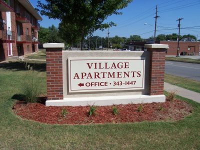 Stone sign outside an apartment complex. It reads village apartments in red letters and there is brick columns on both sides.
